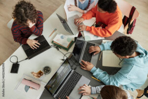 Top view at diverse group of teen school children using computers in classroom studying together at table