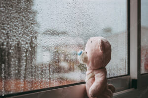 A toy pink sad bear is looking out the window and missing. Autumn rainy day. Raindrops on the window