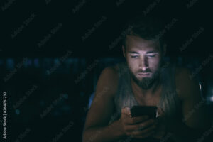 Dark night man looking at mobile phone screen texting late at night awake in bed insomnia or outside in city with skyline background. Serious looking guy depressed addicted to social media.