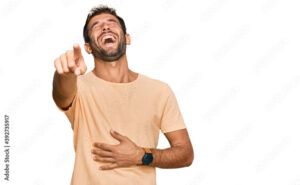Handsome young man with beard wearing casual tshirt laughing at you, pointing finger to the camera with hand over body, shame expression