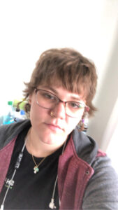 A portrait mode photograph of myself, taken as a selfie. I am wearing a grey and red sweater. I am wearing pink-ish 'clubmaster' eyeglasses. The frame of the photo is slightly angled. I am smiling slightly.