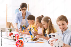 Group of young students in a classroom setting. They are learning about robotics and building their own robots. A teacher is assisting the students, who are all engaged in the activity.