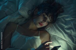 Sleepy exhausted woman lying in bed using smartphone, can not sleep. Insomnia, addiction concept. Sad girl bored in bed scrolling through social networks on mobile phone late at night in dark bedroom