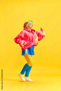 Full-length image of elderly sportive woman in colorful uniform training, running, posing against yellow studio background. Concept of sportive lifestyle, retirement, health care, wellness. Ad