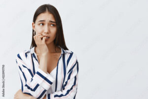 Horizontal shot of anxious good-looking female student in striped blouse, biting fingernail nervously, frowning and looking aside, being scared of consequences, standing over gray background