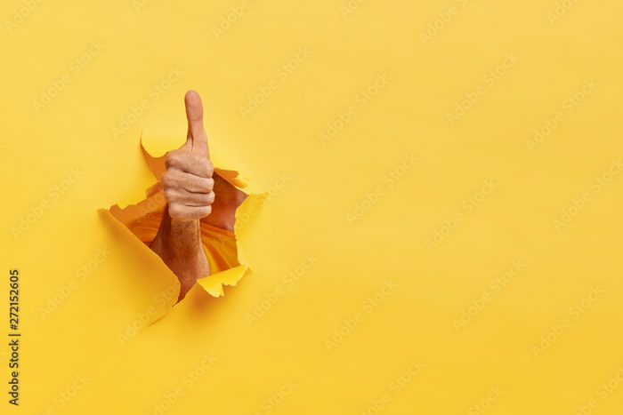 Unrecognizable man shows like gesture through torn yellow wall, keeps thumb up, says you are best, demonstrates approval sign, recommends something. Copy space aside for your advertising content