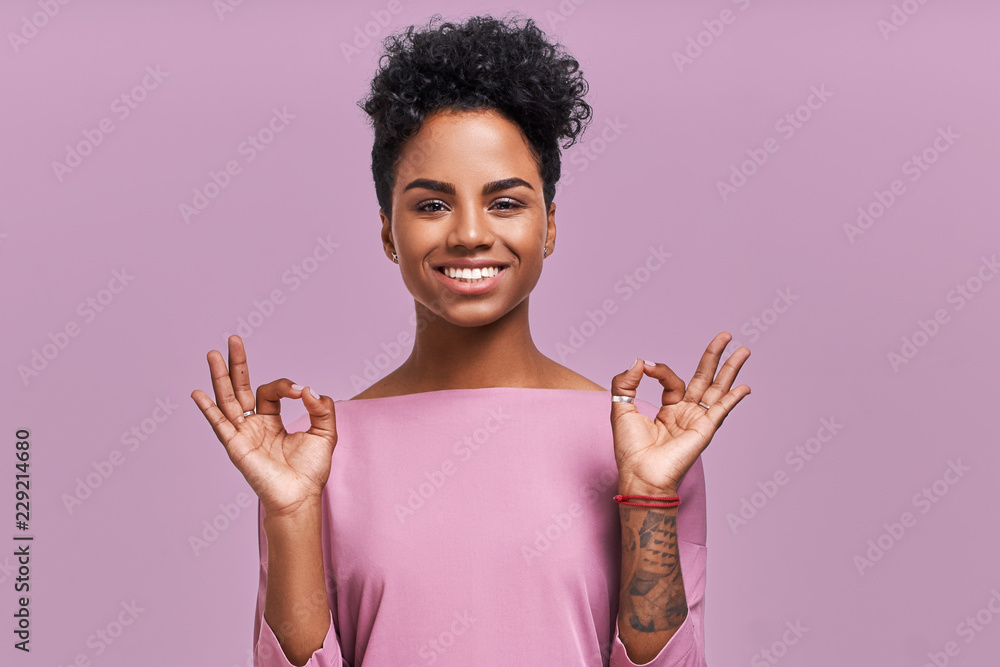 Studio shot of pleased young beautiful African American female shows ok sign, dressed in striped casual jacket, being in good mood, poses against lavender background. People and approval concept
