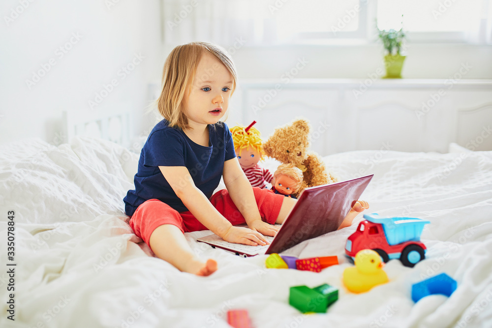 Toddler girl with laptop and toys in bed