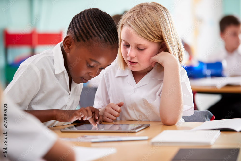 Two Elementary School Pupils Wearing Uniform Using Digital Tablet At Desk