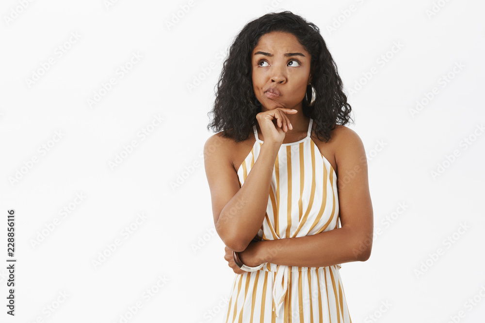 Unsure smart and creative attractive african american female in trendy striped yellow overalls smirking holding hand on chin in thoughtful pose gazing at upper left corner, thinking making choice