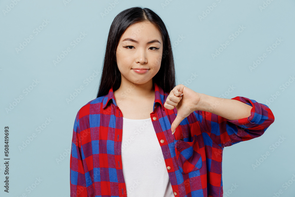 Young sad dissatisfied displeased woman of Asian ethnicity 20s wear checkered shirt showing thumb down dislike gesture isolated on plain pastel light blue color background. People lifestyle concept.