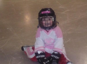 Young child sitting on ice. They wear a hockey helmet, hockey gloves, a pink and white Oilers jersey, and red, pink, and white pants. 