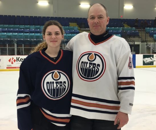 Background: Hockey rink from center ice. Girl on the left, wearing a dark blue Oilers jersey. Man on the right in a white oilers jersey