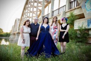 Background: Bridge with graffiti over a river. Five people stand in the foreground. Left to right: Woman in knee-length white dress, Man in blue dress shift, girl in sapphire grad dress, girl in white dress with red flowers. Lastly, girl in a black knee-length dress.