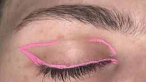 Close up of closed eye. Pink eyeliner on upper lash line. Makes a wing with a line at the end of the wing going about halfway into the lid. Small curved line near top of the inner corner. 