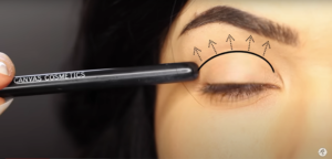 Screenshot of woman with her eye closed. She points end of makeup brush at her eye, which shows the crease with arrows and a line.