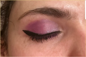 Photo of a closed eye. The eye makeup is a light purple outer corner and crease with a shimmer purple on the inner corners. There is a small black wing along the lash line
