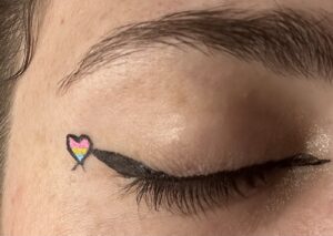 Close-up of an eye. Small winged liner with a heart at the end. Heart is outlined in black with a flag inside. Colours from the top down: Pink, yellow, blue.