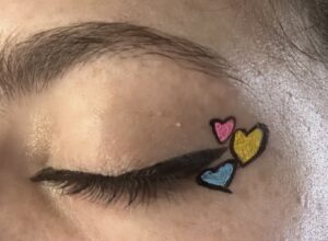 Close up of a closed eye. Small, black winged liner hugging the water line. Three black outlined hearts. One is pink, another is blue, and the last is yellow.