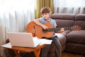 Kid boy playing guitar and watching online lessons on laptop while practicing at home. Stay home. quarantine. Online training, online classes.