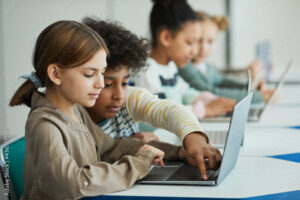 Side view at diverse group of children sitting in row at school classroom and using laptops