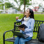 Young female student sitting on a park bench, taking a break from studying with her laptop and notebook, thoughtfully looking up with a pen in hand and headphones around her neck