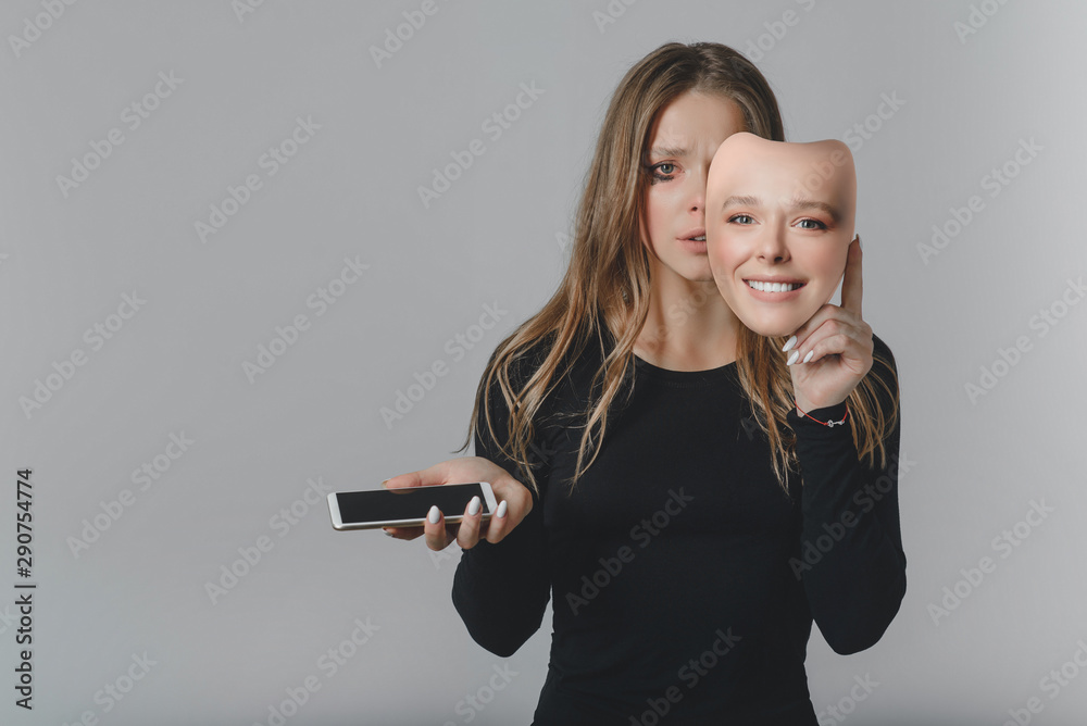 Portrait of a sad girl with a smartphone in one hand and a smiling mask in the other hand.