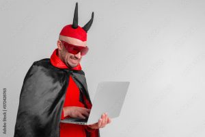 Stylish caucasian man in devil hat with horns and vampire cape with laptop isolated on white backhround.