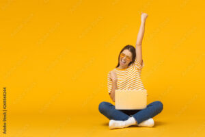 Winner! Excited smiling girl sitting on floor with laptop, raising one hand in the air is she wins, isolated on yellow background