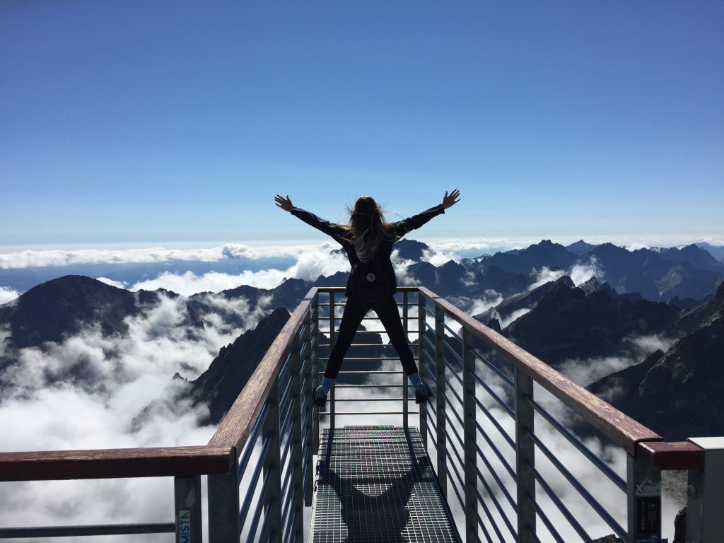 Person standing on hand rails with arms wide open facing the mountains and clouds