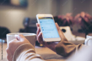 A woman's hands holding white coffee cup and mobile phone while using social media in modern loft cafe