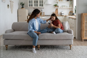 Caring attentive mother stroking daughter sits on couch with phone in hands offers to take walk and spend time together. Kind woman works as nanny looking after teenage girl during absence of parents