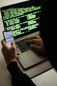 Unrecognizable hacker with smartphone typing on laptop at desk