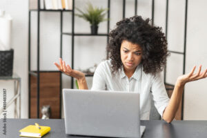Young African American female feeling stressed, tired, and confused, sitting at the desk in front of laptop, struggling and raising hands up