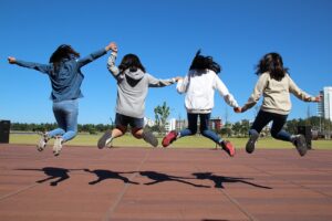 A photo of four youth, holding hands, jumping into the air. They are facing away from the camera. The sky is blue suggesting a sunny fall day. Their shadows reflect onto the playground.