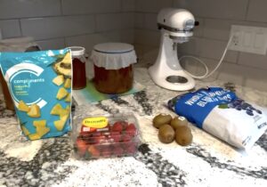 A bag of frozen pineapple, a package of strawberries, four kiwis, and a bag of frozen whole blueberries on a kitchen counter with kombucha and a mixer in the background.