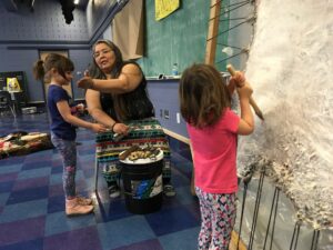 two children and female Indigenous knowledge keeper scraping buffalo hide and smudging