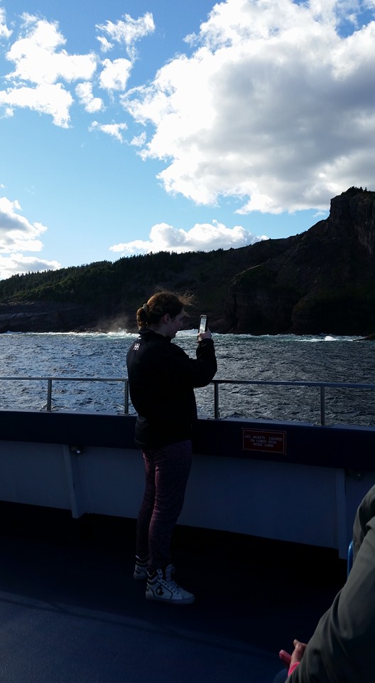 Background: Coastline of Newfoundland. Foreground: Girl stands on a boat taking photos of the coast. 