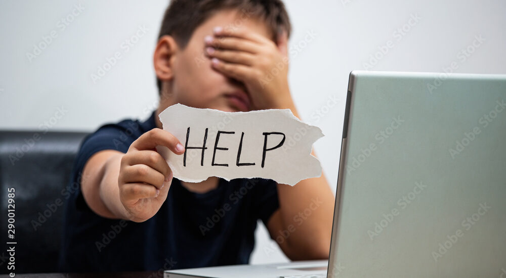 Sad and scared young boy with computer laptop suffering cyberbullying and harassment being online abused by stalker or gossip feeling desperate and humiliated in cyber bullying concept.