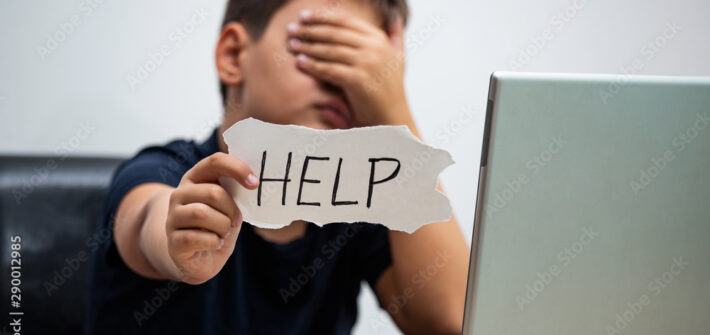 Sad and scared young boy with computer laptop suffering cyberbullying and harassment being online abused by stalker or gossip feeling desperate and humiliated in cyber bullying concept.