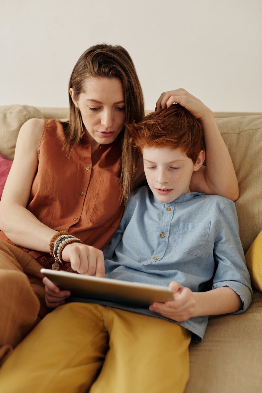 photo of woman and boy sitting on couch while using tablet computer